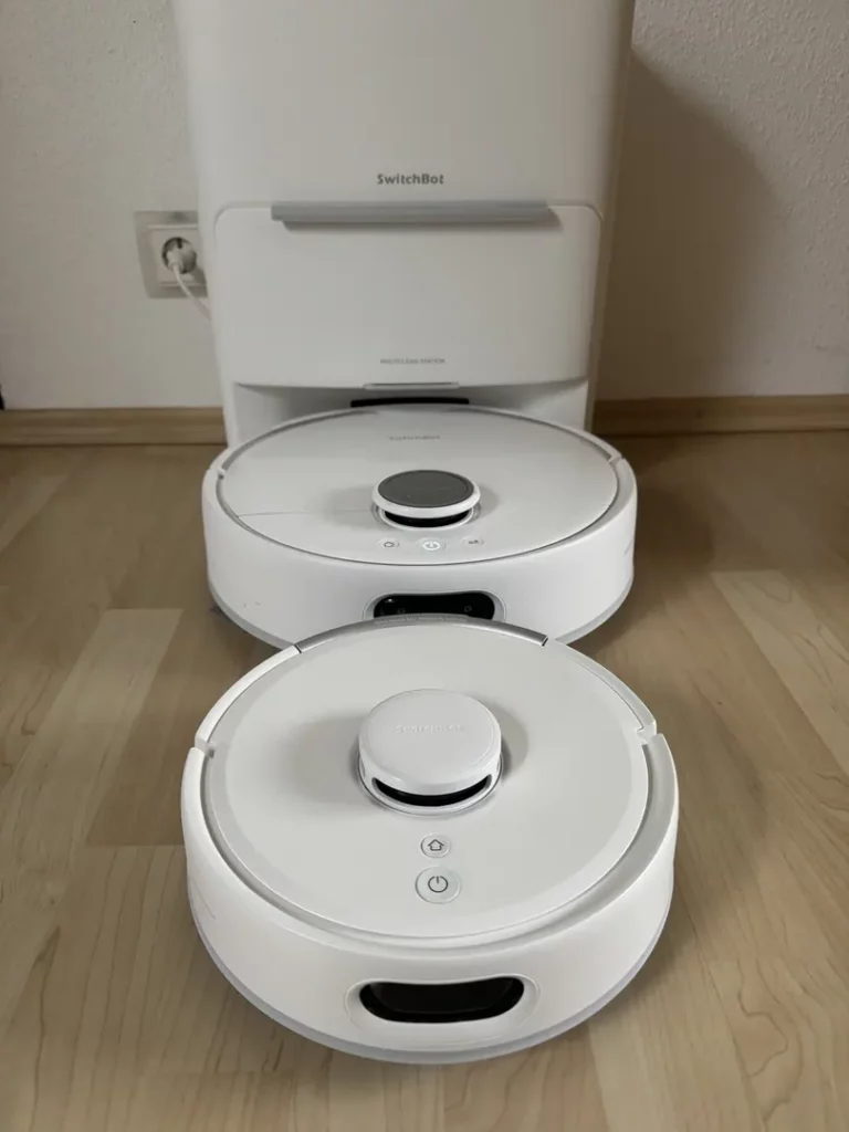 Zwei weiße Saugroboter stehen nebeneinander auf einem hellen Holzboden, während im Hintergrund eine weiße Basisstation zu sehen ist; die Geräte haben runde Formen und sind mit Tasten zur Steuerung ausgestattet.