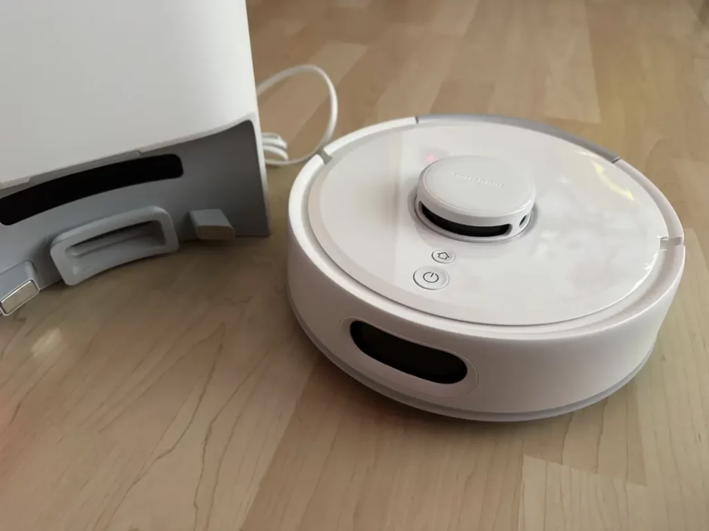 Weißer Roboterstaubsauger auf einem hellen Holzboden, neben einem grauen Ladegerät, zeigt moderne Technologie und Gerätekonstruktion.