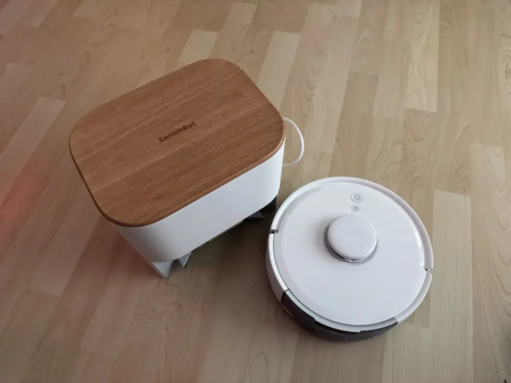 Weißer Saugroboter und ein Smart-Home-Gerät mit Holzoberfläche auf einem hellen Holzboden, ideal für moderne Wohnräume.