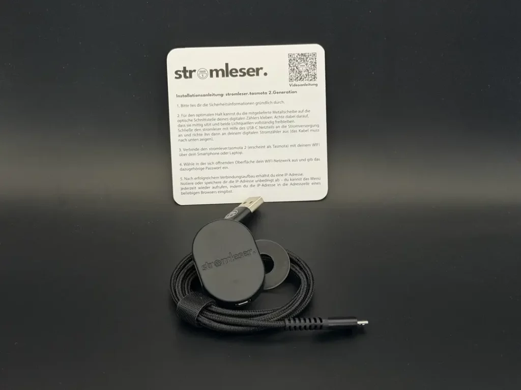 Ein schwarzes, rundes Gerät mit dem Schriftzug „stromleser“ und einem USB-C-Anschluss liegt auf einem dunklen Hintergrund. Neben dem Gerät befindet sich ein Informationsblatt mit Anweisungen zur Installation des stromlesers sowie einem QR-Code für weitere Informationen. Das Kabel des Geräts ist ebenfalls zu sehen.