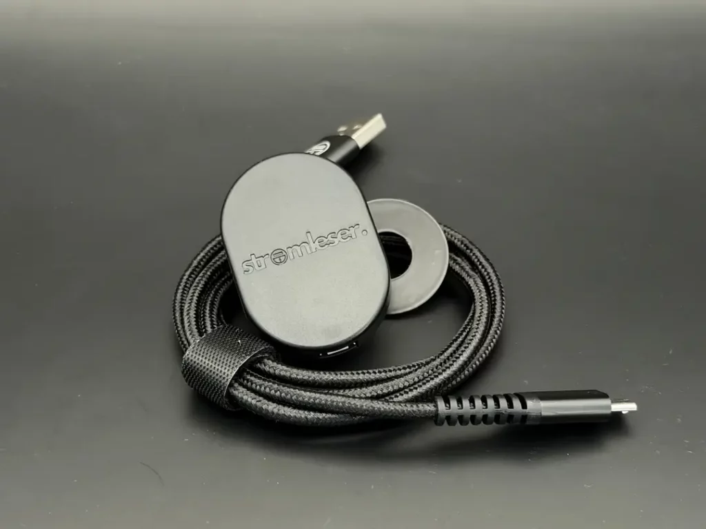 USB-C Kabel mit einem schwarzen, ovalen Adapter und einem runden, grauen Ring, alles auf einem schwarzen Hintergrund angeordnet.