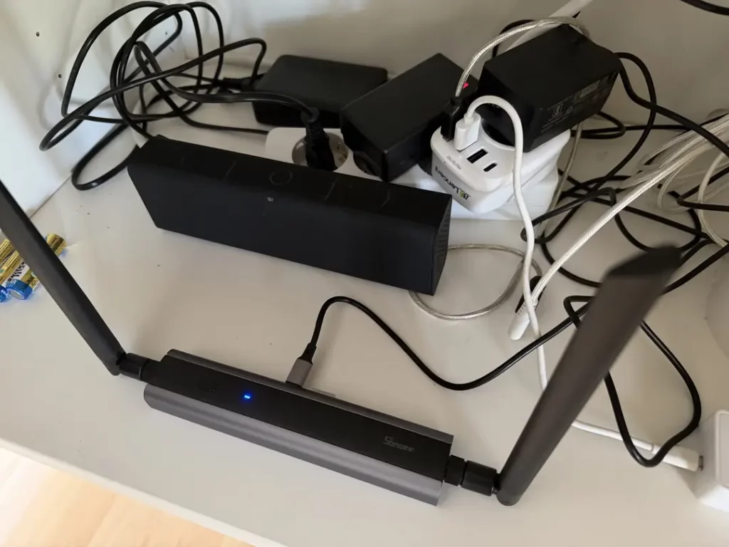 Ein unordentliches Kabelmanagement mit einem WLAN-Router im Vordergrund, der zwei Antennen hat und durch ein blaues Licht leuchtet. Im Hintergrund sind verschiedene elektronische Geräte zu sehen, darunter ein Lautsprecher, ein Netzteil und mehrere aufgerollte Kabel, die lose um die anderen Geräte herumliegen.