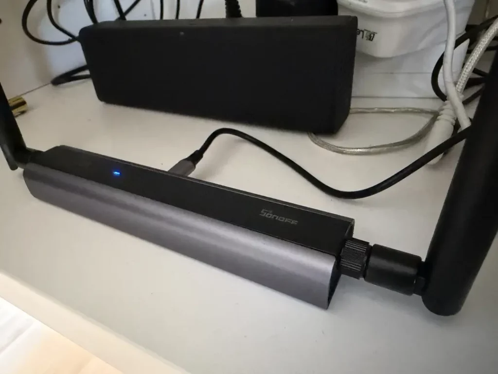 Ein moderner Sonos Verstärker in schwarzem und silbernem Design, auf einem weißen Regal platziert, mit angeschlossenen Kabeln und einem blauen LED-Licht, das seinen Betriebsstatus anzeigt, umgeben von weiteren elektronischen Geräten.