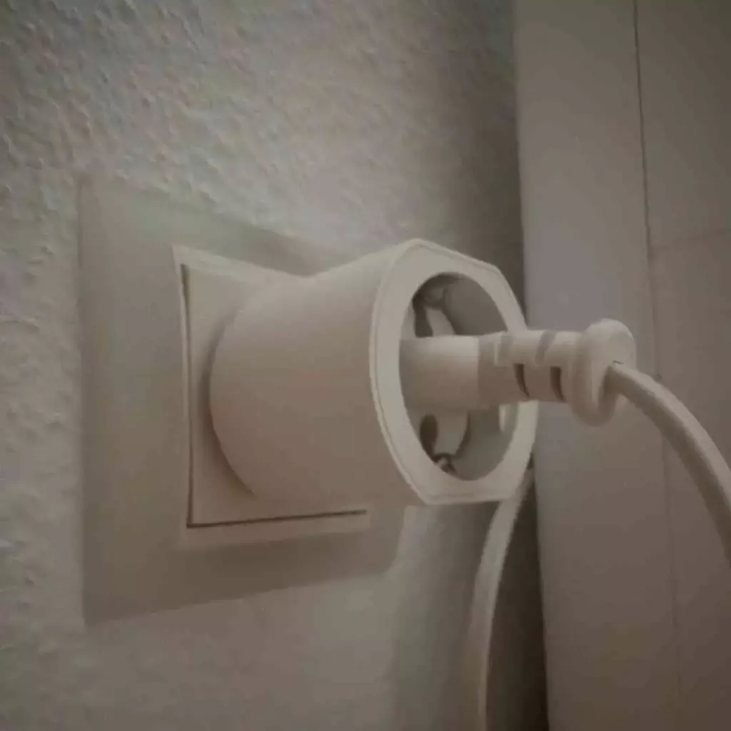 Steckdose mit einem weißen Stecker, der teilweise eingesteckt ist, an einer hellen Wand, neben einem Kabel, das aus der Steckdose herausführt.