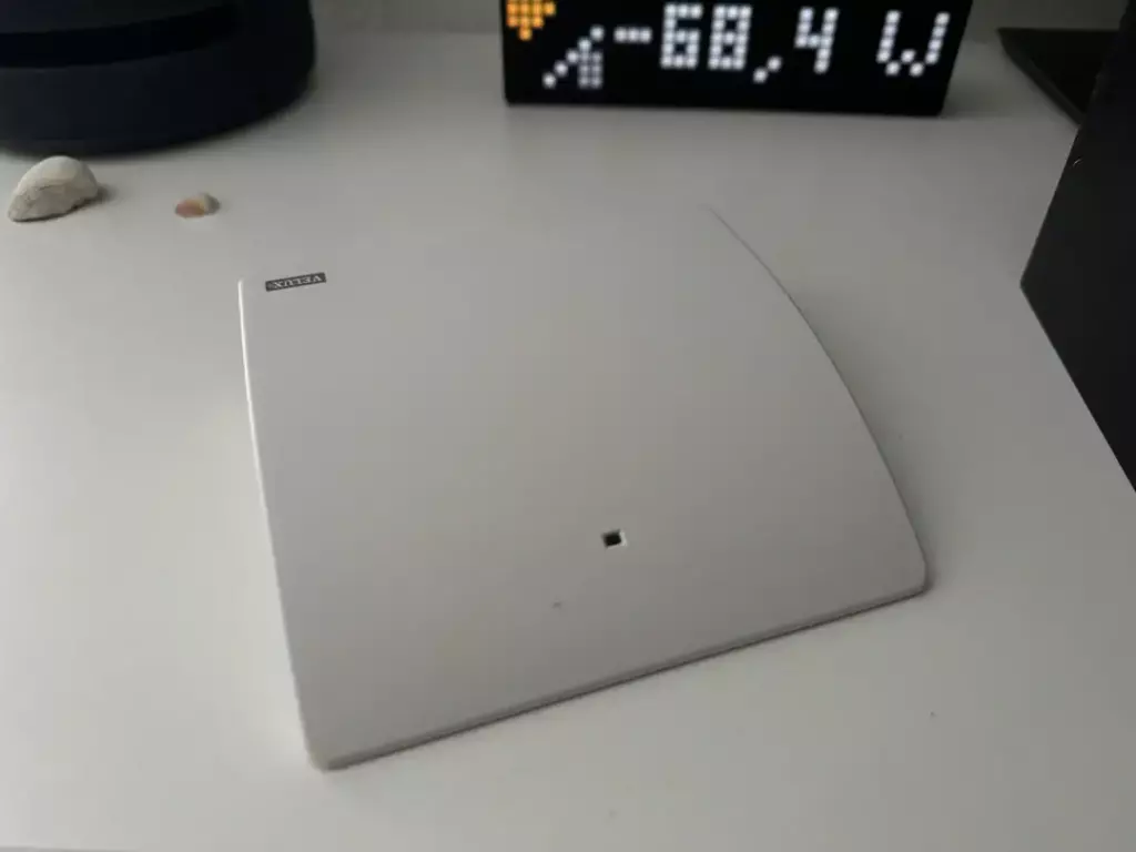 Ein quadratisches, weißes Gerät auf einem Tisch, das wahrscheinlich ein Smart Home Sensor oder Schalter ist, mit einer kleinen Erhebung in der Mitte; im Hintergrund ein digitaler Zähler, der eine Leistungsanzeige in Watt (68,4 W) zeigt.